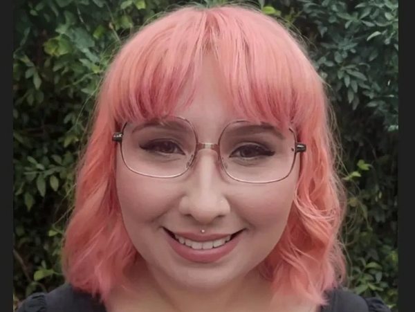 Smiling woman with pink hair and glasses outdoors.