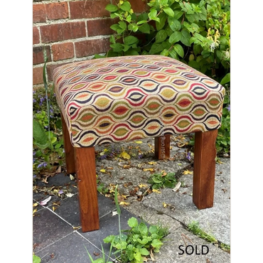 Wooden stool with colorful patterned upholstery, placed outdoors against a brick wall.