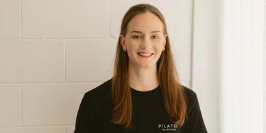 Smiling woman in black Pilates t-shirt standing against a white brick wall.
