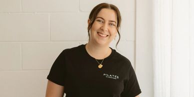 Smiling woman in black Pilates shirt standing near a white curtain.