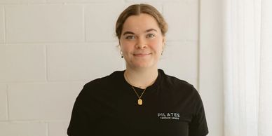 Young woman in a black Pilates T-shirt standing in front of a white brick wall.