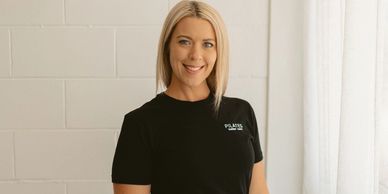 Smiling woman in black Pilates outfit standing by a white curtain and wall.