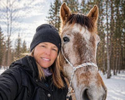 A woman in winter clothing takes a selfie with a horse in a snowy forest.