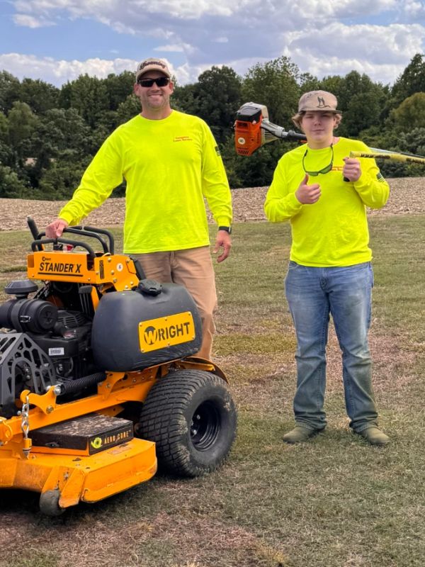 Two men in bright yellow shirts with lawn care equipment outdoors.