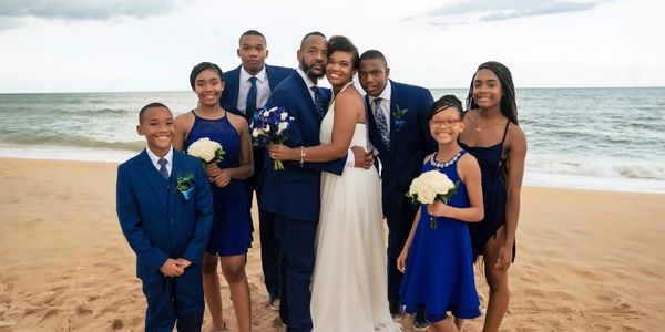 A joyful family dressed in blue and white poses on the beach at a wedding.
