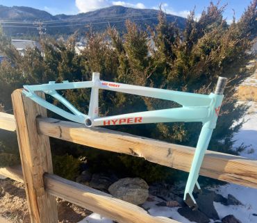 Light blue Hyper bicycle frame hanging on wooden fence outdoors.