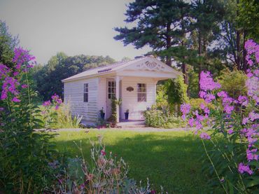 Small white cottage surrounded by greenery and pink flowers.