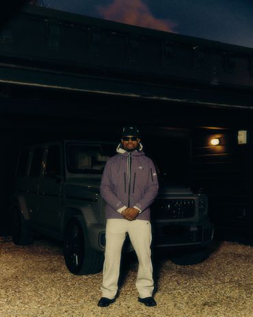 Man in a jacket and cap standing in front of a dark SUV at night.