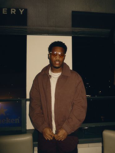 A man wearing a brown jacket and glasses posing indoors at night.