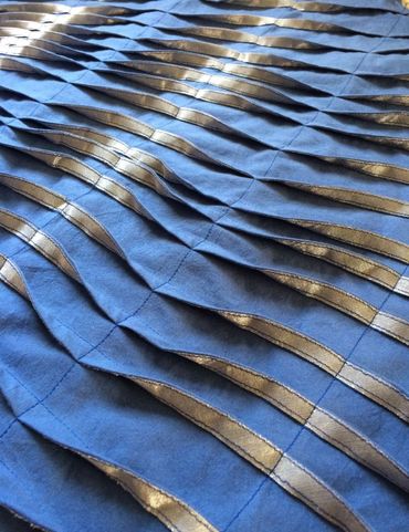 Detail image of pleated and machine stitched  blue fabric with silver applique.