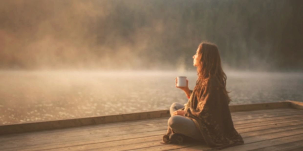 Woman enjoying a warm drink by a misty lake at sunrise.