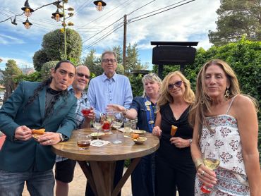 Group of six people enjoying drinks outdoors around a wooden table.