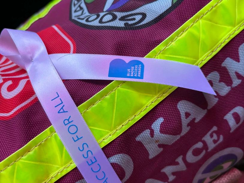 A purple ribbon with 'ACCESS FOR ALL' and Blue Badge Access Awards text on a colorful fabric background.