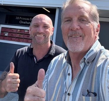 Two men smiling and giving thumbs up outdoors.