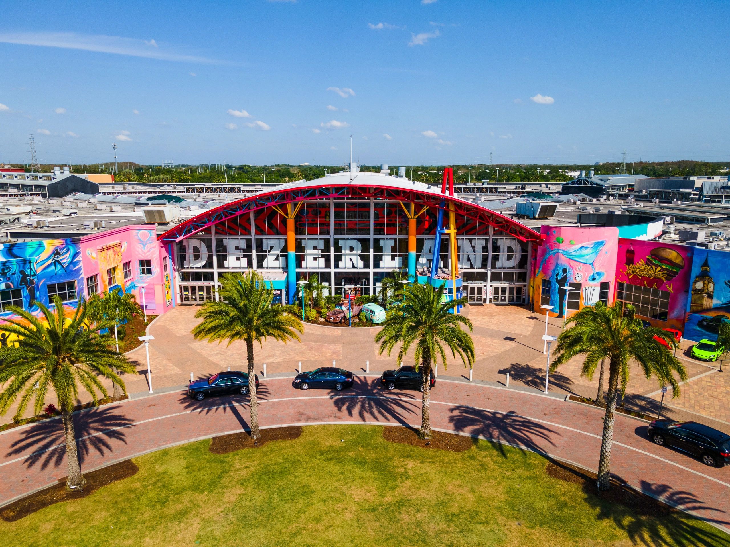 Colorful entrance of Dezerland with palm trees and cars in front.