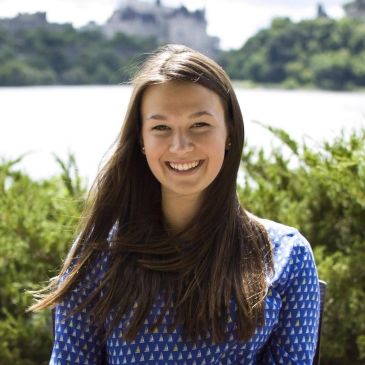 Young woman smiling outdoors with greenery and water in the background.