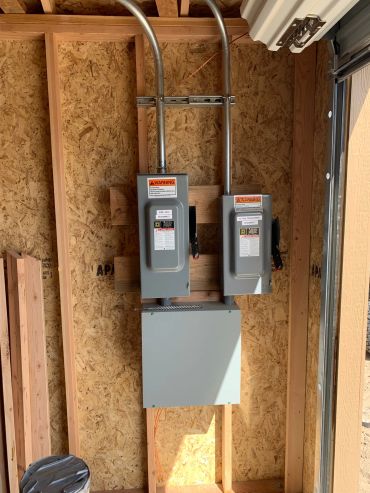 Two electrical panels mounted on a wooden wall in a garage.