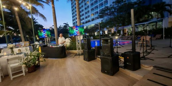 Outdoor DJ setup with speakers and screens near a pool at dusk.