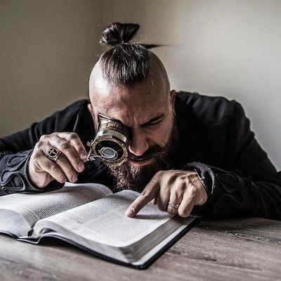 A man with a top knot uses a monocle to read a book intensely.