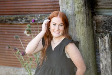 Young woman with red hair smiles by a wooden post.