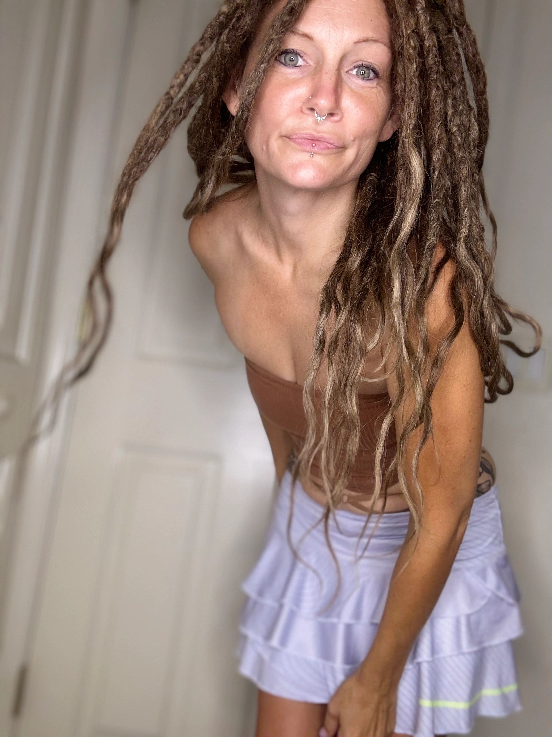 Woman with dreadlocks and piercing wearing a brown top and white skirt.