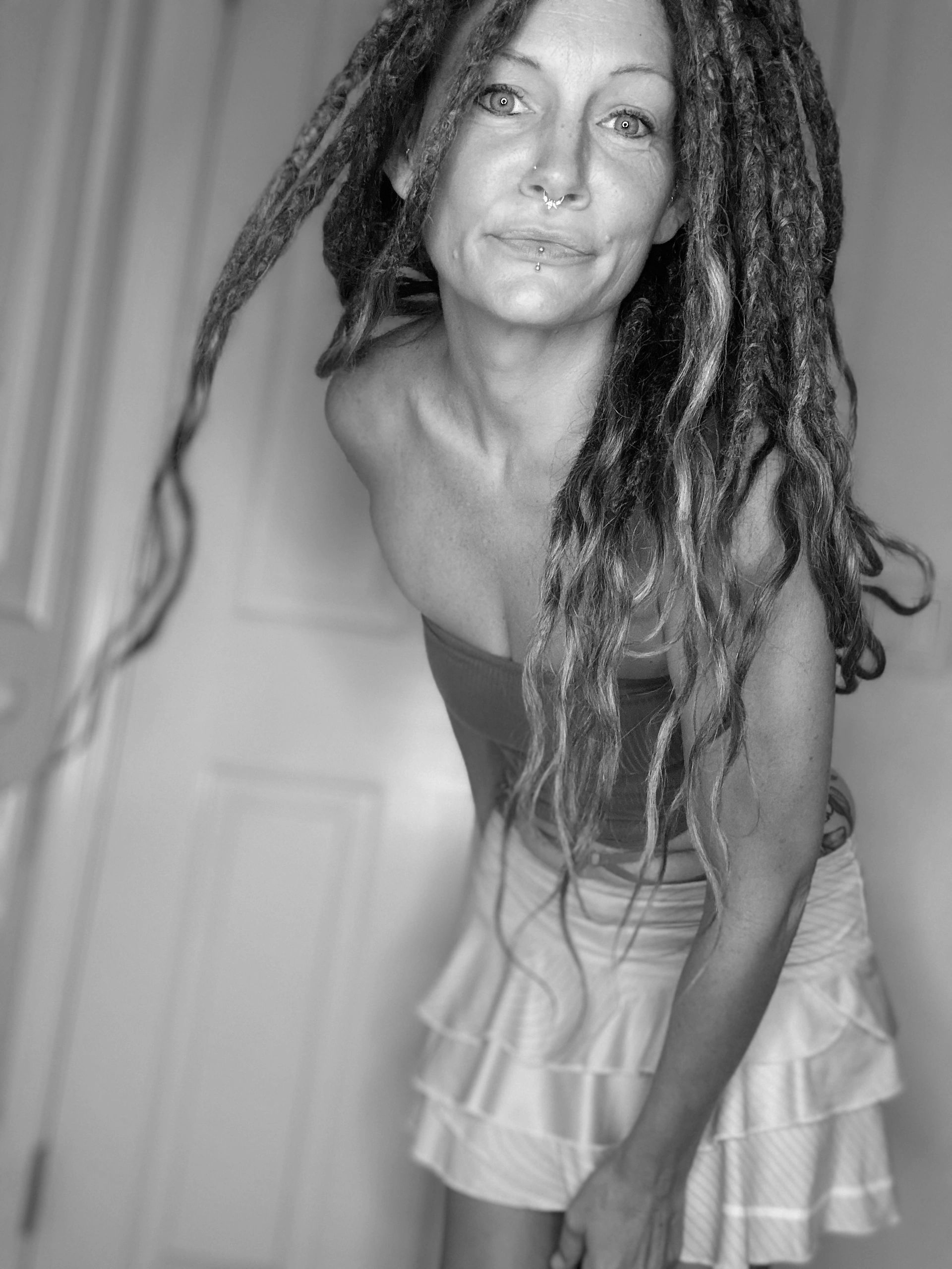 Woman with dreadlocks, piercings, and a layered skirt leaning forward.