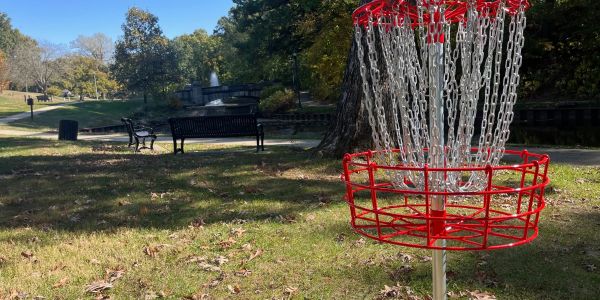 A red disc golf basket in a park with benches and trees in the background.