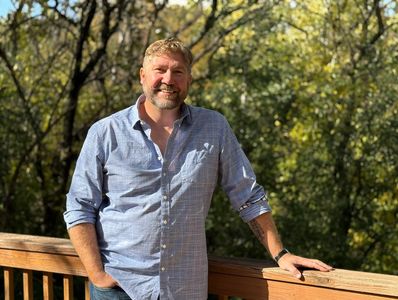 Man leaning on wooden railing outdoors with trees in background.