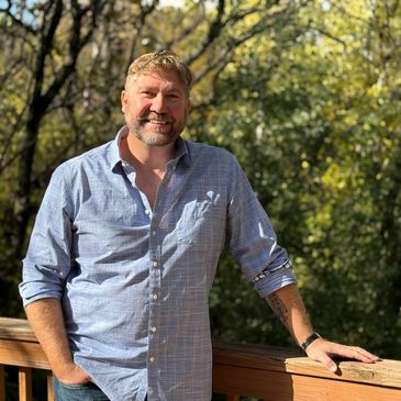 Man leaning on wooden railing outdoors with trees in background.