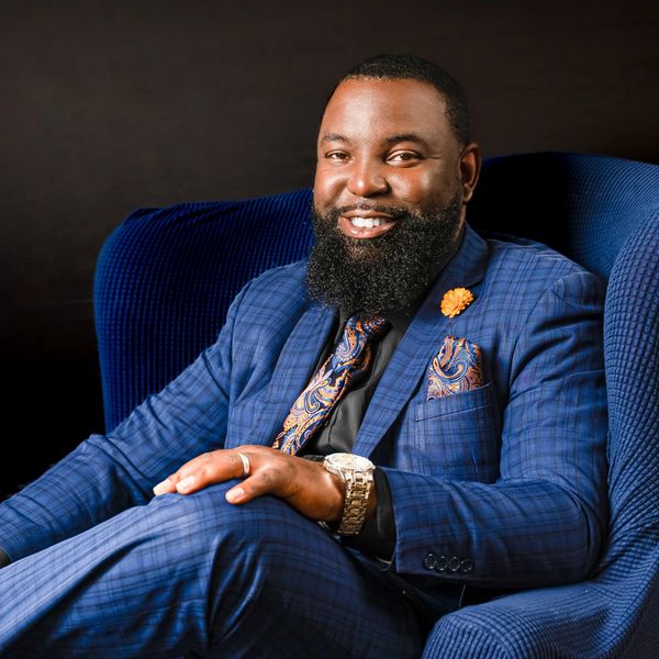 Man in a blue suit smiling while sitting on a blue chair.