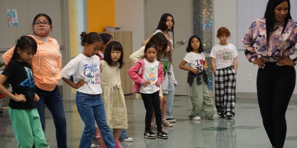 Group of children standing in a line with a woman in a floral jacket.