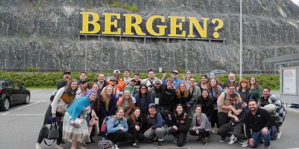 A large group poses in front of a rock face with 'BERGEN?' sign.