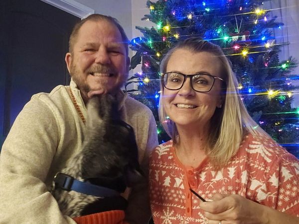A joyful couple and their dog in festive attire by a Christmas tree.