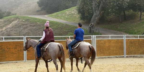 Two people riding horses in an outdoor arena with a hilly background.