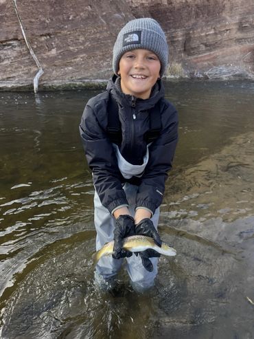 Smiling boy holding a fish in a river, dressed for fishing.