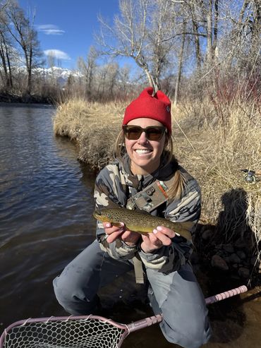 Smiling woman in red beanie holding a fish by the river.