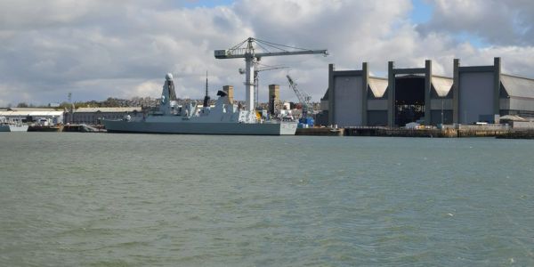 Naval ship docked at a harbor with cranes and buildings.