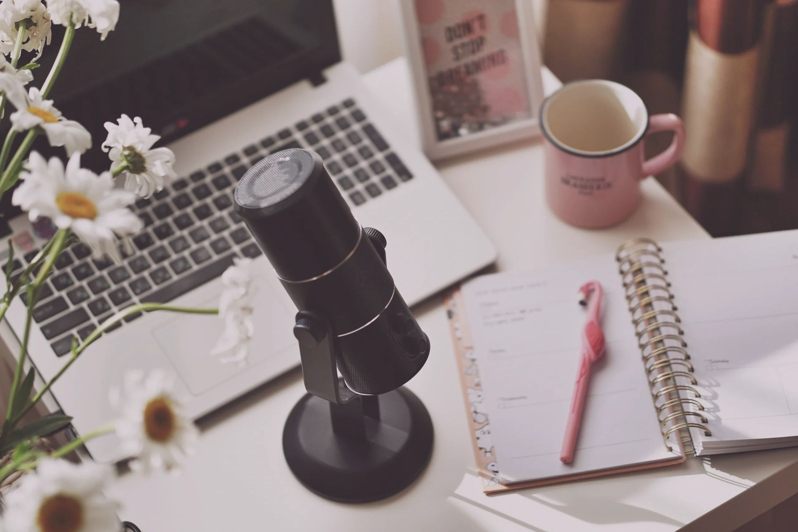 Podcast-Szene: Mikro, Laptop, Kalender, rosa Tasse und Margheriten auf einem Schreibtisch