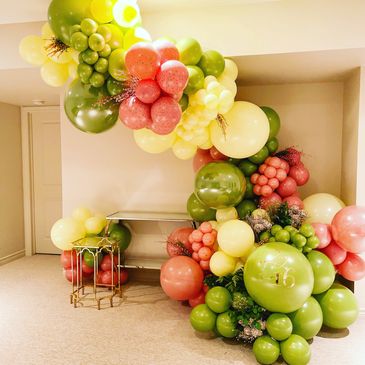 Colorful balloon arch with green, yellow, and pink balloons in a room corner.