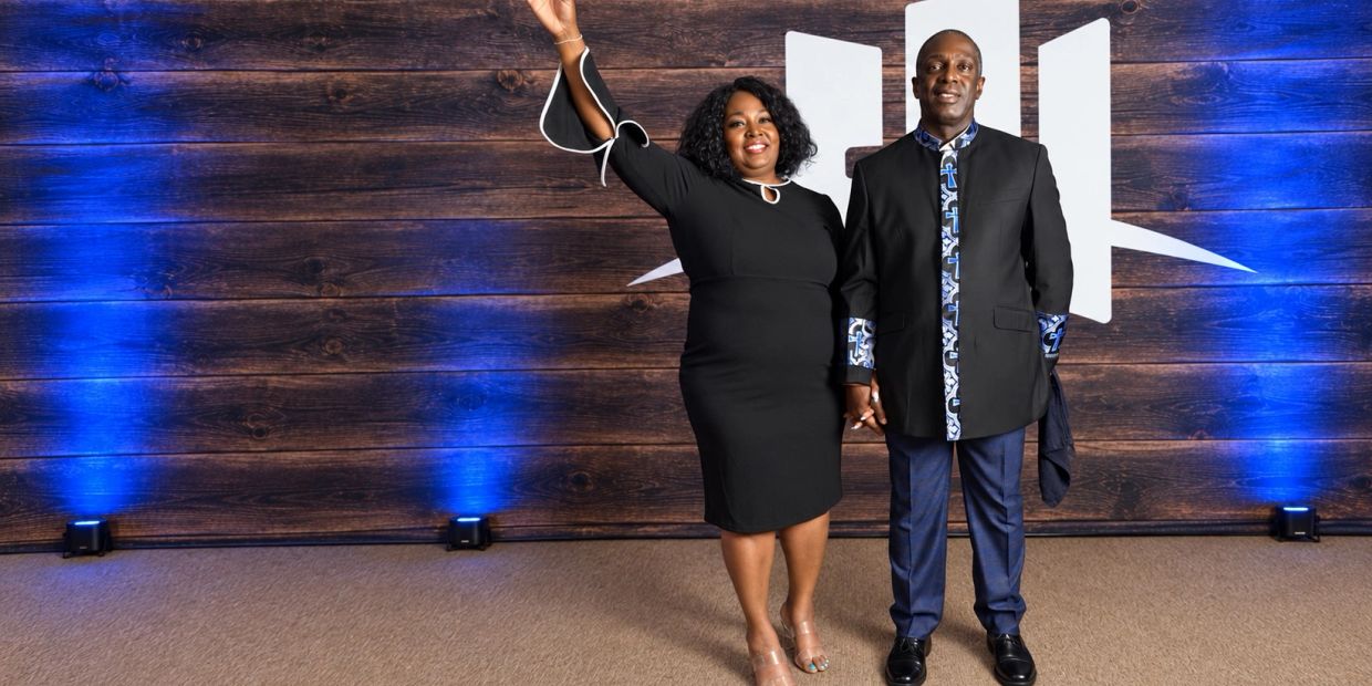 A smiling couple dressed formally stands against a wooden backdrop with blue lights.