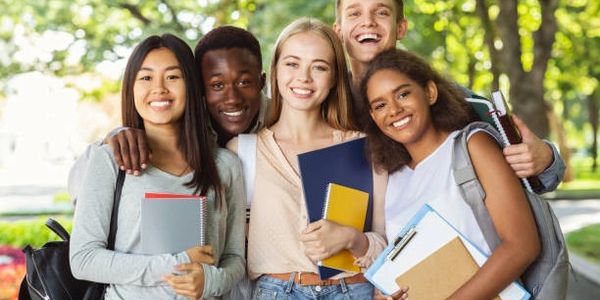 Group of five diverse students smiling outdoors with books and backpacks.