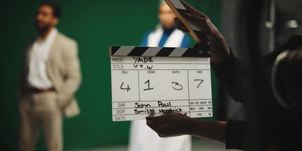 Film clapperboard in focus with actors blurred in the background.