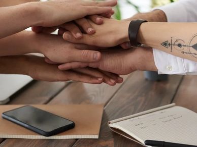 Arms reaching into to stack hands on top of each other. Underneath are phones, notepads, and pens