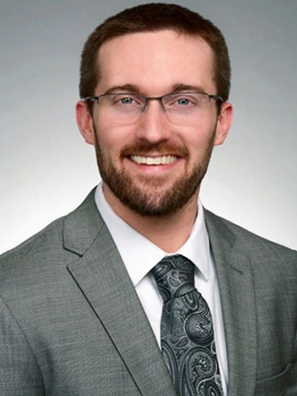 Professional man with glasses in gray suit and patterned tie smiling.