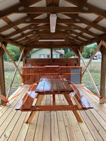 Wooden picnic table and kitchen area under a covered pavilion.