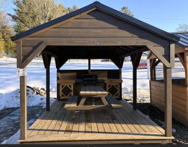 Wooden pavilion with picnic table and grill in snowy outdoor setting.