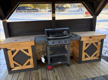 Outdoor gas grill station with wooden storage cabinets on snowy day.