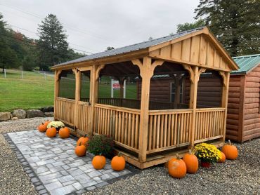 Wooden screened gazebo surrounded by pumpkins and fall flowers on a stone patio.