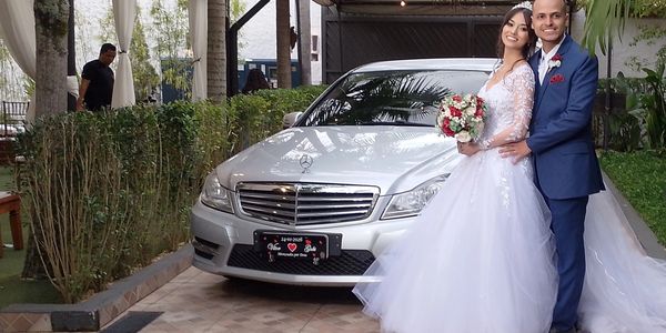 Casal de noivos sorrindo ao lado de um carro prateado.