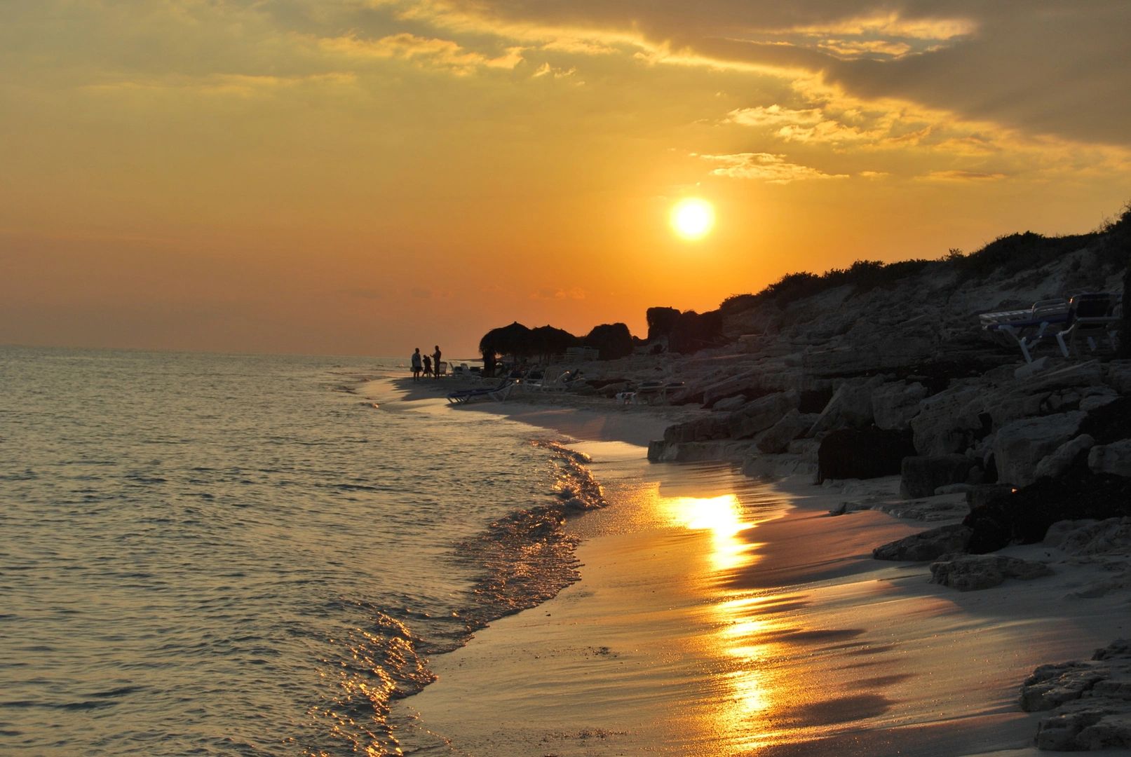 Sunset over a tranquil beach with gentle waves and silhouettes of people.